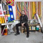 Color photo of Michael Hambouz seated in a studio and surrounded by his artwork