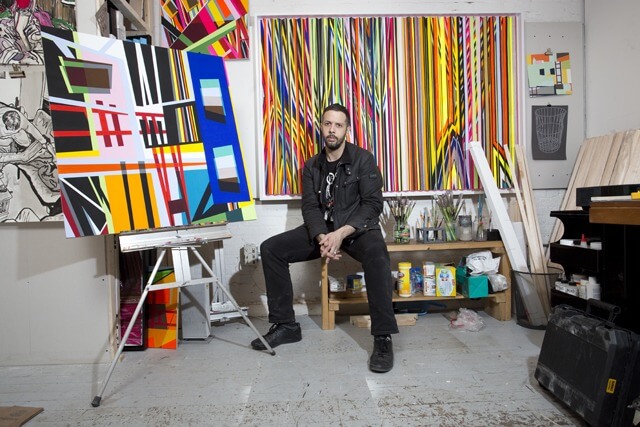 Color photo of Michael Hambouz seated in a studio and surrounded by his artwork