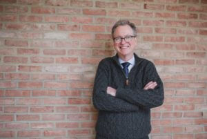 Photo of President Tom Manley standing in front of a wall with his arms crossed
